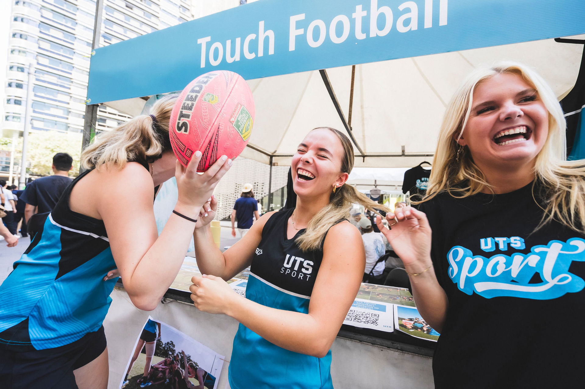 Three female members laughing in front of the UTS Touch Football stall. One student is holding a football.