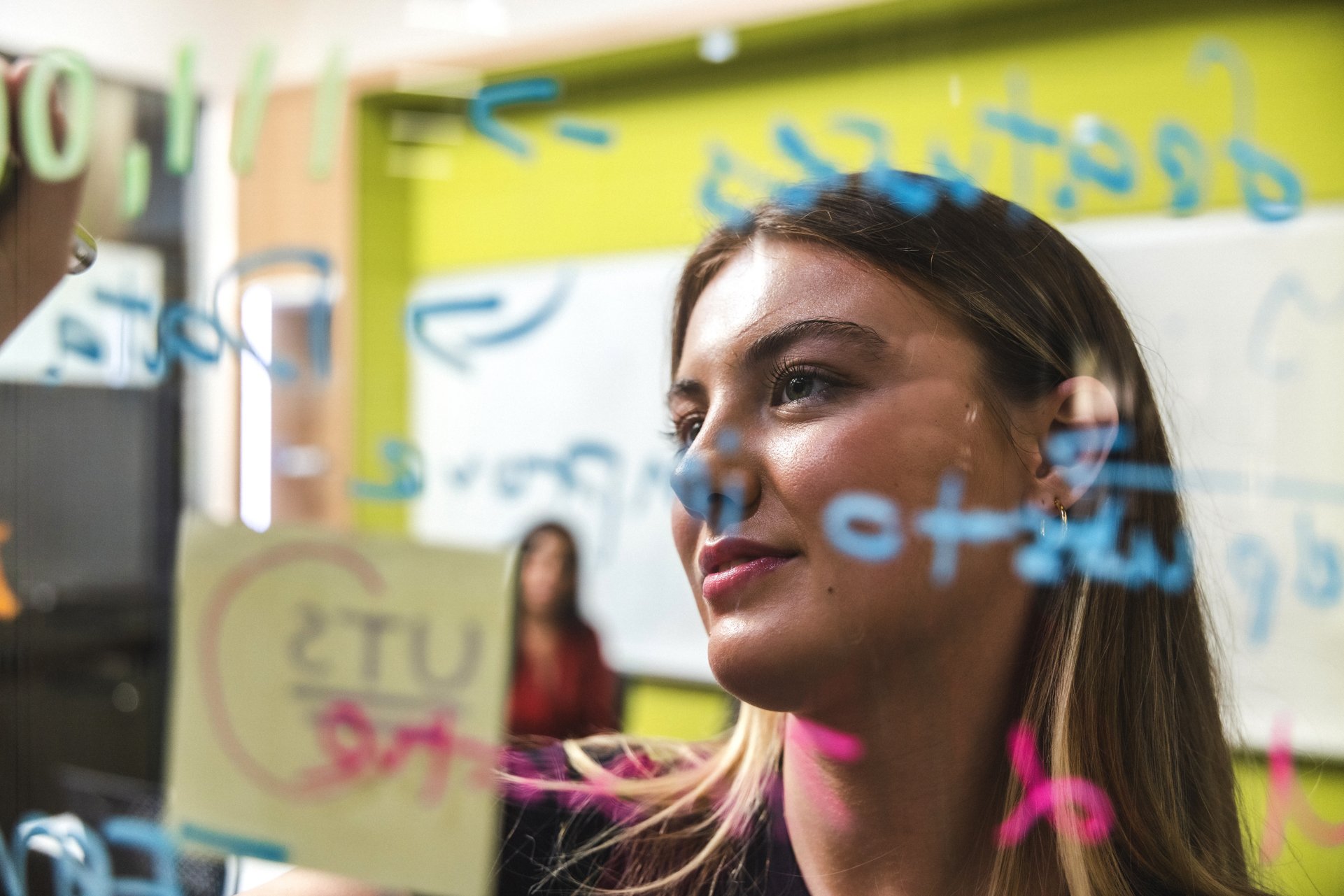 Female student writing on glass wall in office and brainstorming with colleague in the background.